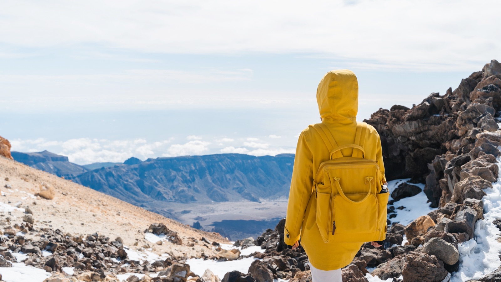 Hiker in a yellow jacket walking a snow-dusted trail high on Mount Teide, with the caldera far below